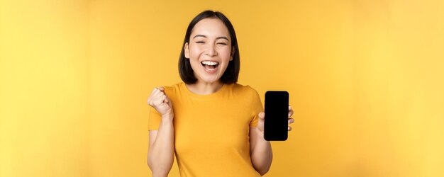 Donna asiatica eccitata che mostra l'app per smartphone e festeggia trionfante sul telefono cellulare in piedi su sfondo giallo Spazio di copia