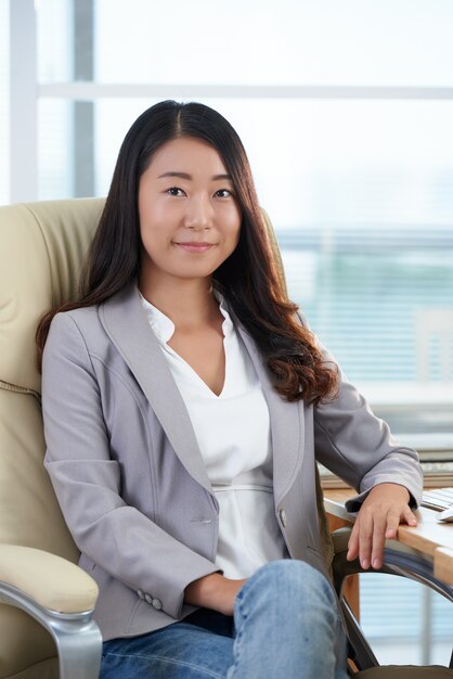 Donna asiatica elegantemente vestita sicura che si siede nella sedia esecutiva in ufficio