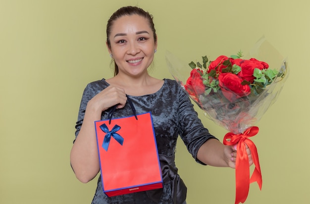 Donna asiatica felice che tiene il mazzo delle rose rosse e del sacchetto di carta con il regalo che sorride allegramente celebrando la giornata internazionale della donna in piedi sopra la parete verde