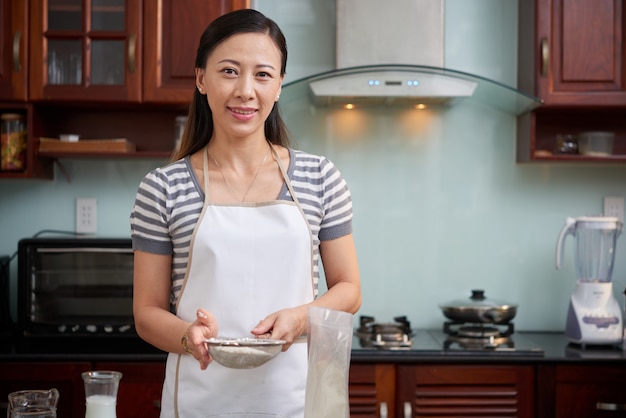 Donna asiatica felice nel setaccio della tenuta del grembiule con farina in cucina a casa