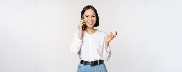 Donna asiatica felice sorridente che parla sullo smartphone con la commessa del cliente su chiamata che tiene il telefono cellulare e che fa gesti in piedi su sfondo bianco