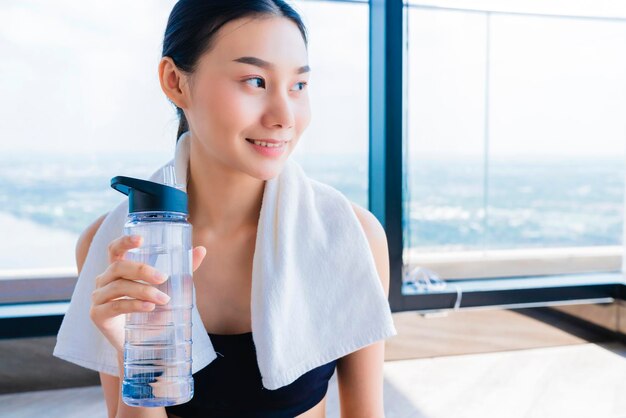 Donna asiatica in buona salute che si rilassa in palestra dopo l'allenamento tiene la mano della bevanda dell'acqua pura e dell'asciugamano bianco