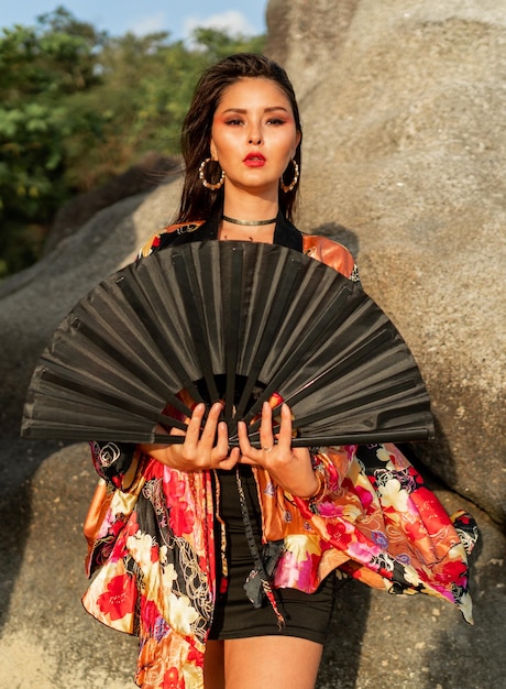 Donna asiatica in camicetta kimono di seta colorata che tiene ventaglio nero e posa sulla spiaggia con rocce