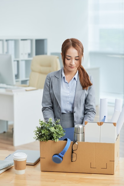 Donna asiatica in vestito che sta nell'ufficio con gli effetti personali in scatola di cartone sullo scrittorio