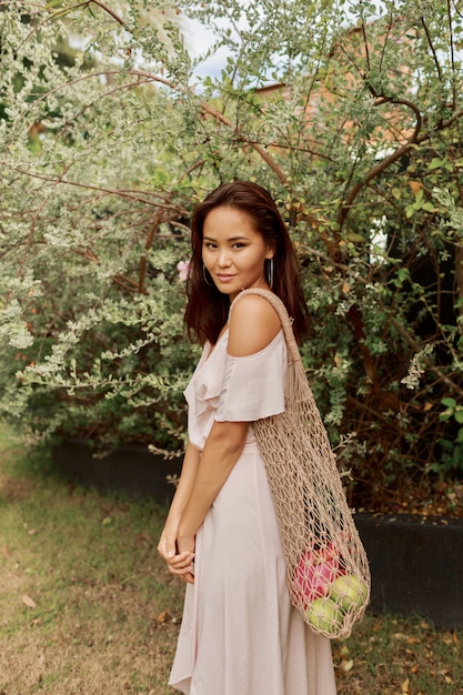 Donna asiatica in vestito che tiene la borsa shopper amichevole della maglia di eco con i frutti tropicali freschi.