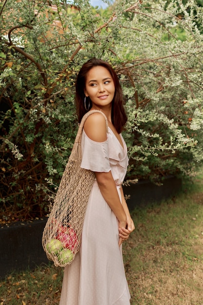 Donna asiatica in vestito che tiene la borsa shopper amichevole della maglia di eco con i frutti tropicali freschi.