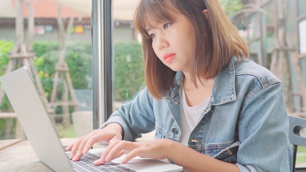 Donna asiatica indipendente di affari che lavora, facendo progetti e inviando email sul computer portatile o sul computer mentre sedendosi sulla tavola in caffè.
