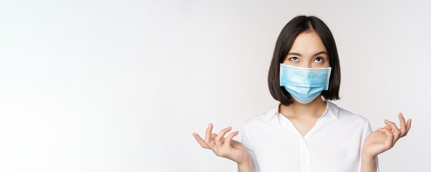 Donna asiatica infastidita in maschera medica che scrolla le spalle e alza lo sguardo con l'espressione del viso infastidita stan