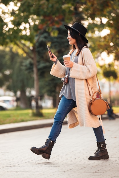 Donna asiatica moderna che utilizza smartphone e che tiene caffè asportabile mentre camminando nel parco all'aperto