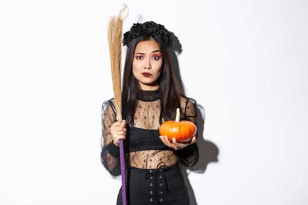 Donna asiatica preoccupata e confusa in costume da strega che sembra nervosa, tenendo in mano scopa e zucca, dolcetto o scherzetto su Halloween, in piedi su sfondo bianco.