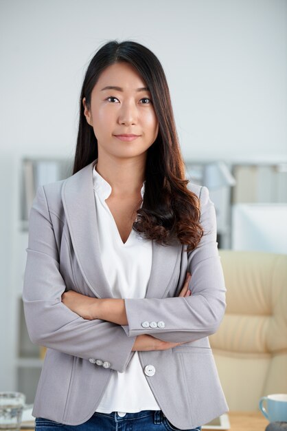 Donna asiatica sicura in rivestimento astuto che propone nell'ufficio con le braccia attraversate