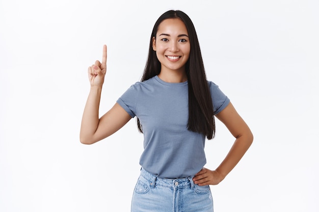Donna asiatica sorridente allegra e spensierata bruna in maglietta casual, tenere la mano sulla vita con un atteggiamento positivo che punta il dito verso l'alto, mostrandone uno, sorridendo, dare consigli su cosa comprare, promuovere il prodotto