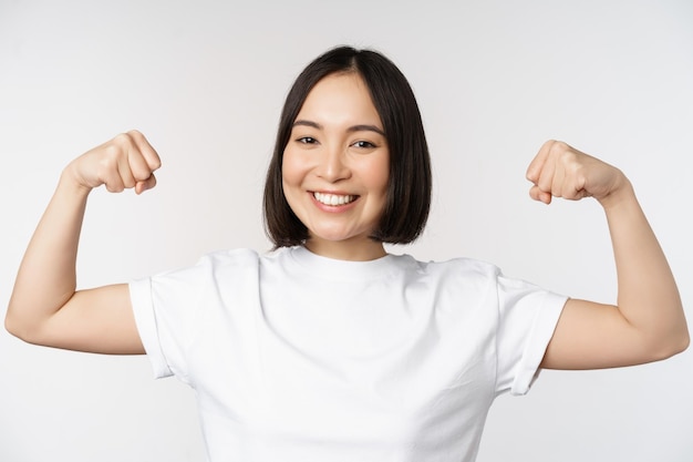 Donna asiatica sorridente che mostra la flessione dei muscoli del bicipite forte gesto delle braccia in piedi in maglietta bianca su sfondo bianco