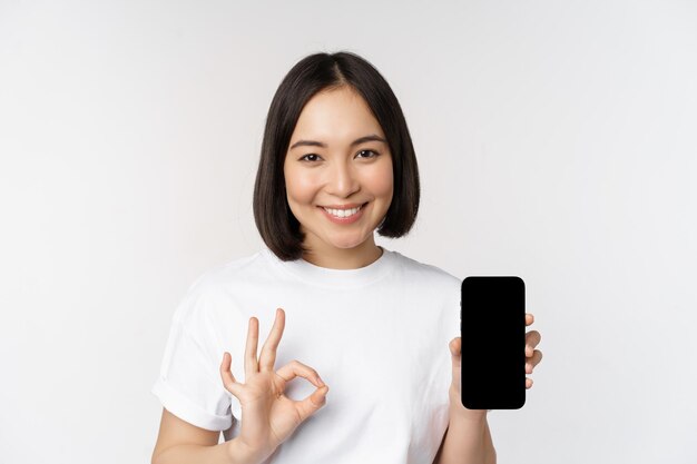 Donna asiatica sorridente che mostra lo schermo del telefono cellulare ok consigliando l'app per smartphone in piedi su sfondo bianco