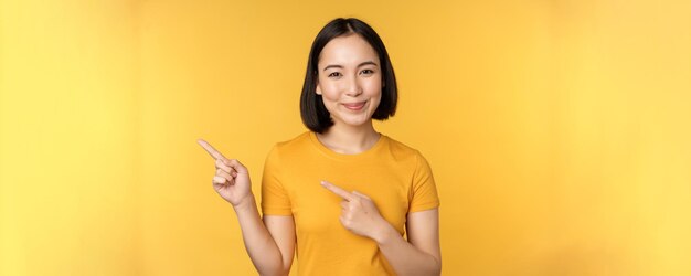 Donna asiatica sorridente che punta le dita a sinistra mostrando pubblicità su uno spazio vuoto di copia in piedi su sfondo giallo