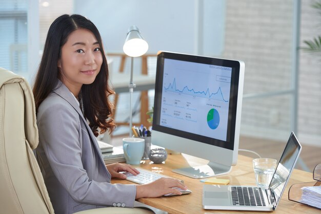 Donna asiatica sorridente che si siede allo scrittorio in ufficio e che lavora al rapporto finanziario
