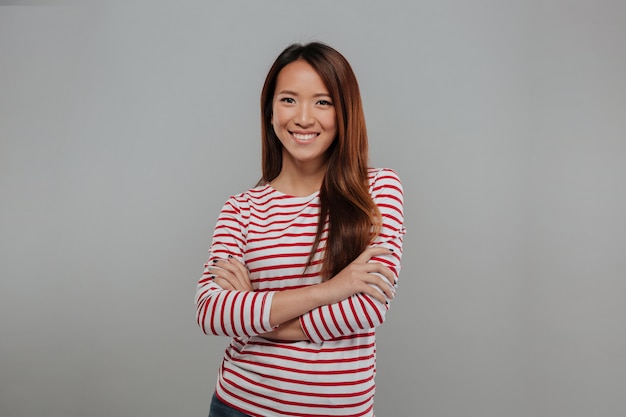 Donna asiatica sorridente in maglione che posa con le armi attraversate e che esamina la macchina fotografica sopra fondo grigio