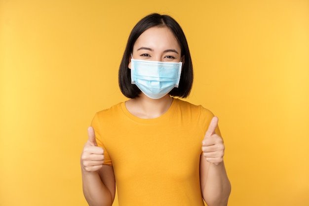 Donna asiatica sorridente in maschera medica che mostra i pollici in su come approvazione e raccomanda smth in piedi su sfondo giallo