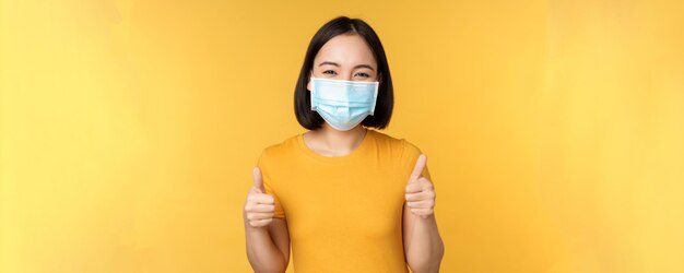 Donna asiatica sorridente in maschera medica che mostra i pollici in su come approvazione e raccomanda smth in piedi su sfondo giallo