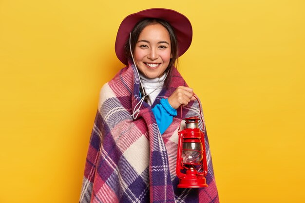 Donna asiatica sorridente soddisfatta avvolta in un plaid, tiene una piccola lanterna a gas, indossa un cappello, posa su sfondo giallo studio