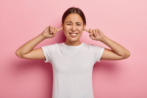 Donna asiatica stressata infastidita da un forte rumore, tappi le orecchie con gli indici, evita suoni sgradevoli, si acciglia insoddisfatta, ha i capelli scuri, indossa una maglietta bianca, modelle contro il muro rosa