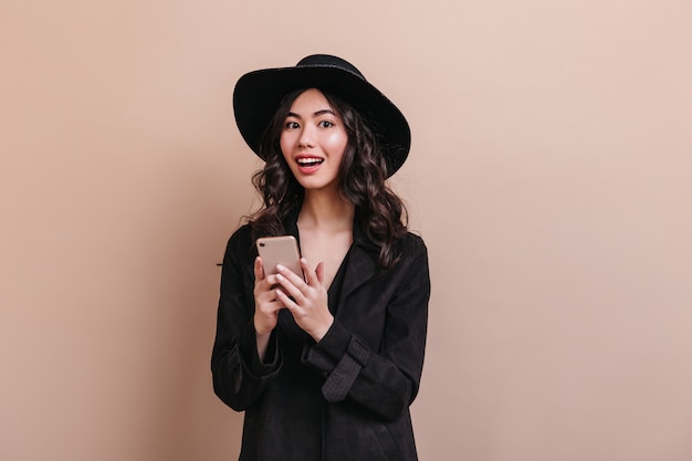 Donna asiatica stupita che tiene smartphone e che guarda l'obbiettivo. Elegante donna riccia in cappotto in posa con gadget.