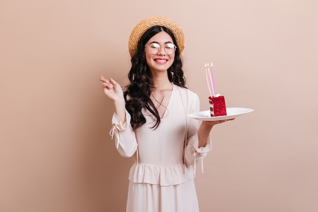 Donna asiatica sveglia che ride mentre fa il desiderio di compleanno. Raffinata donna giapponese con cappello tenendo un pezzo di torta.