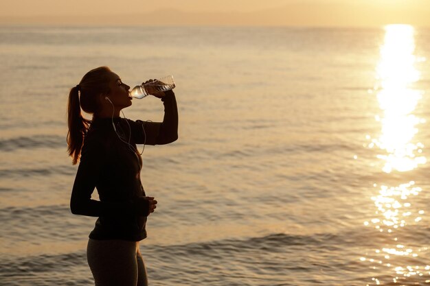 Donna atletica assetata che beve acqua da una bottiglia e si rinfresca in riva al mare all'alba Spazio di copia