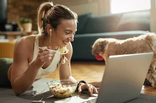 Donna atletica che si diverte mentre mangia insalata e naviga in rete sul computer portatile.