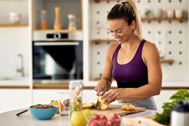 Donna atletica felice che taglia la frutta mentre prepara un pasto sano in cucina