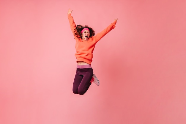 Donna attiva in felpa con cappuccio arancione e leggings scuri che salta vigorosamente sul muro rosa