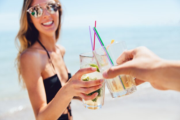 Donna attraente allegra in occhiali da sole che beve un cocktail con amico, tostatura