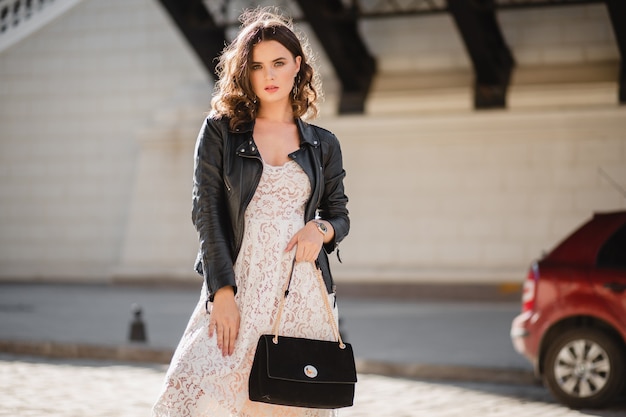 Donna attraente che cammina per strada in abito alla moda, tenendo la borsa, guardando verso il basso, indossa una giacca di pelle nera e abito di pizzo bianco, stile primavera