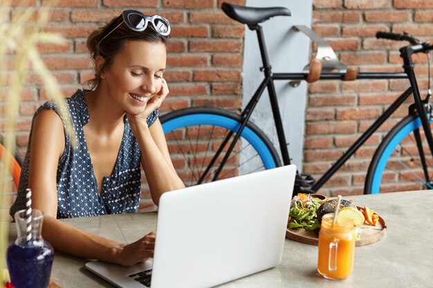 Donna attraente che indossa occhiali da sole sulla testa facendo videochiamata al suo ragazzo, sorridendo timidamente, appoggiandosi con il gomito sul tavolo al caffè. Amici di messaggistica femminile carino online