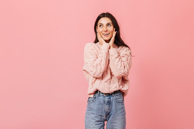 Donna attraente con gli occhi azzurri sorride dolcemente su sfondo rosa. Signora in pullover lavorato a maglia tocca il suo viso.