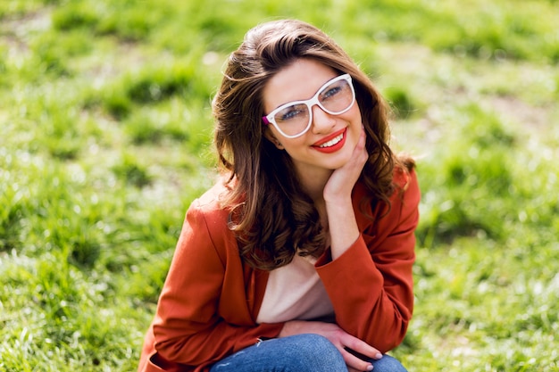 Donna attraente con labbra carnose, occhiali da vista, giacca rossa, acconciatura ondulata che si siede sull'erba verde nel parco soleggiato di primavera e sorridente