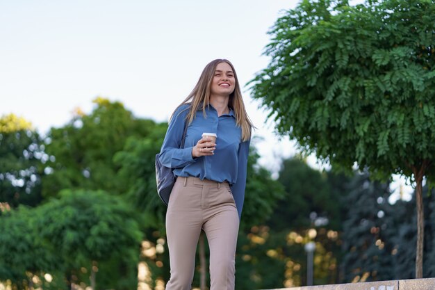 Donna attraente del primo piano in movimento con caffè da asporto sulla strada della città. Ragazza bionda del ritratto che tiene tazza di carta con la bevanda calda all'aperto.