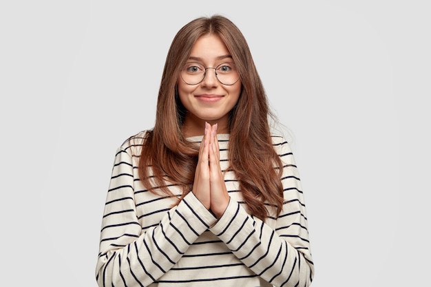 Donna attraente e soddisfatta con un sorriso gentile, tiene le mani in preghiera, vestita con abiti a righe