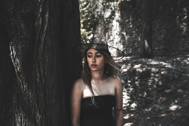 Donna attraente in cappello da strega accanto all&#39;albero e guardando lontano