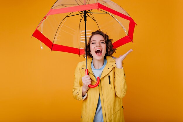 Donna attraente in cappotto giallo di autunno che esprime emozioni positive. Raffinata ragazza con i capelli ricci corti che ride sotto l'ombrello.