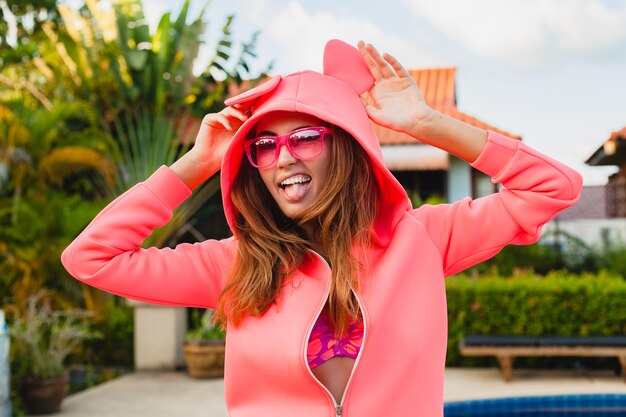 Donna attraente in felpa con cappuccio rosa colorato che indossa occhiali da sole in vacanza estiva sorridente espressione emotiva del viso divertendosi, stile di moda sportiva