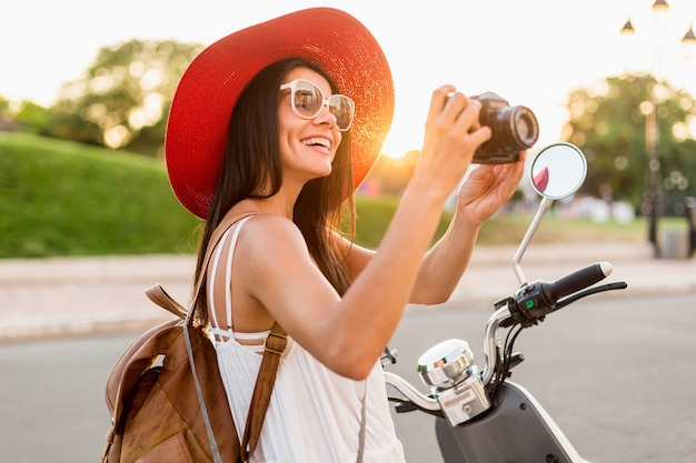 Donna attraente in sella a una moto in strada, stile di vacanza estiva, viaggiare, sorridere, divertirsi, abbigliamento elegante, avventure, scattare foto con la macchina fotografica vintage, indossare lo zaino in pelle