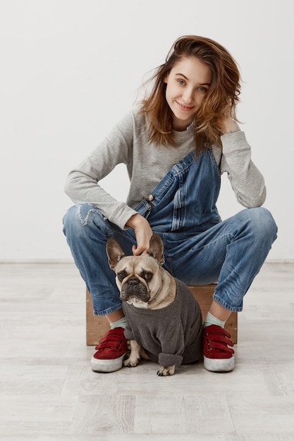 Donna attraente in tuta di jeans che riposa a casa con un cucciolo adorabile che ha emozioni piacevoli