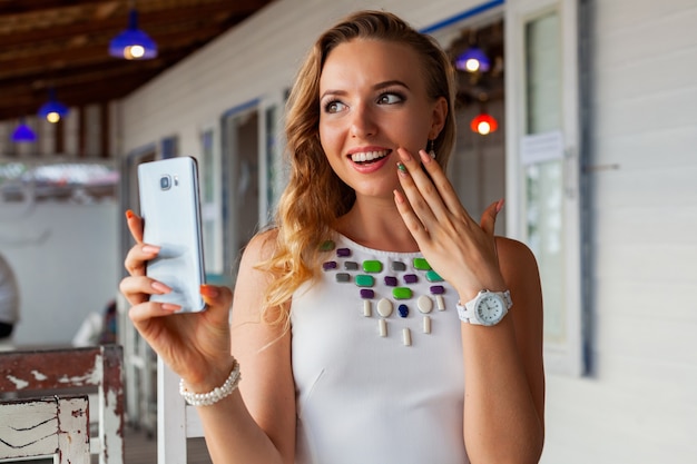 Donna attraente in vestito bianco in caffè estivo in occhiali da sole utilizzando il telefono che cattura foto