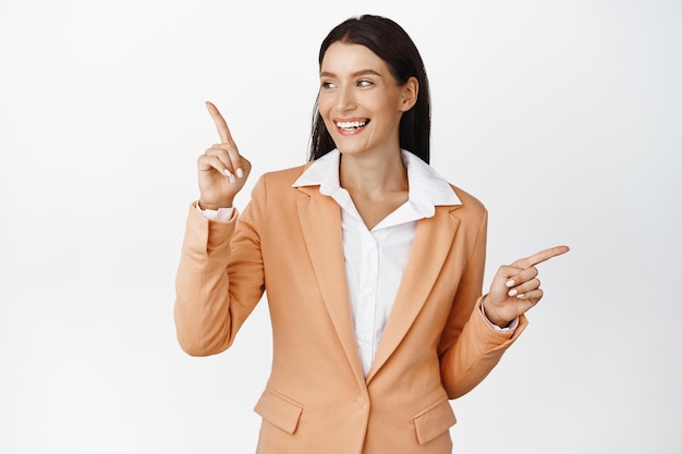 Donna aziendale di successo in tuta sorridente che punta le dita lateralmente guardando a sinistra la pubblicità della società del logo in piedi su sfondo bianco