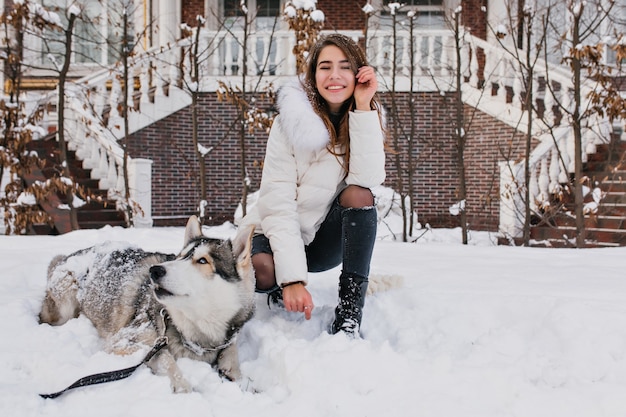 Donna bianca con un sorriso incredibile in posa con il suo cane durante la passeggiata invernale in cortile. La foto all'aperto della signora allegra indossa pantaloni di jeans strappati seduti sulla neve con un husky pigro.