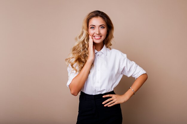 Donna bionda adorabile con il sorriso perfetto in blusa bianca che posa sopra la parete beige. Abito elegante da lavoro.