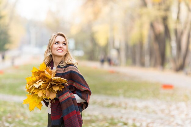 Donna bionda allegra che cammina nel parco autunnale con un bouquet di foglie d'acero