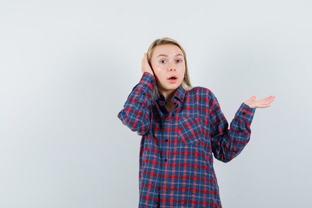 Donna bionda alzando la mano mentre si preme la mano sull'orecchio, che propone alla macchina fotografica in camicia controllata e guardando sorpreso. vista frontale.