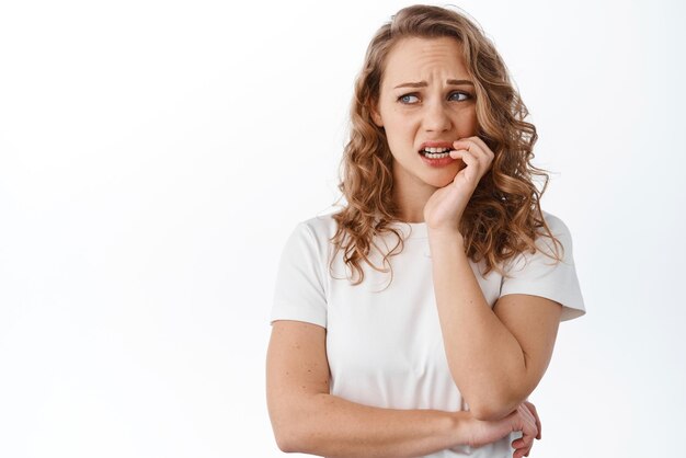 Donna bionda ansiosa che si morde le unghie e guarda da parte con la faccia nervosa spaventata in piedi nervosa su sfondo bianco